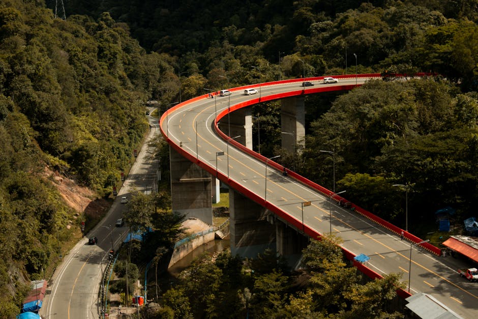 Winding highway with red railings cutting through a lush green mountainous area, showcasing modern infrastructure.